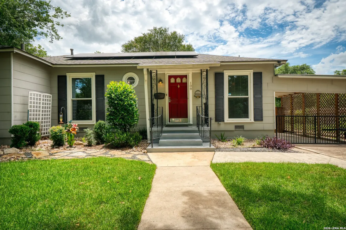 Single-story gray home with a bright red front door, black shutters, solar panels, and a concrete walkway under blue sky.