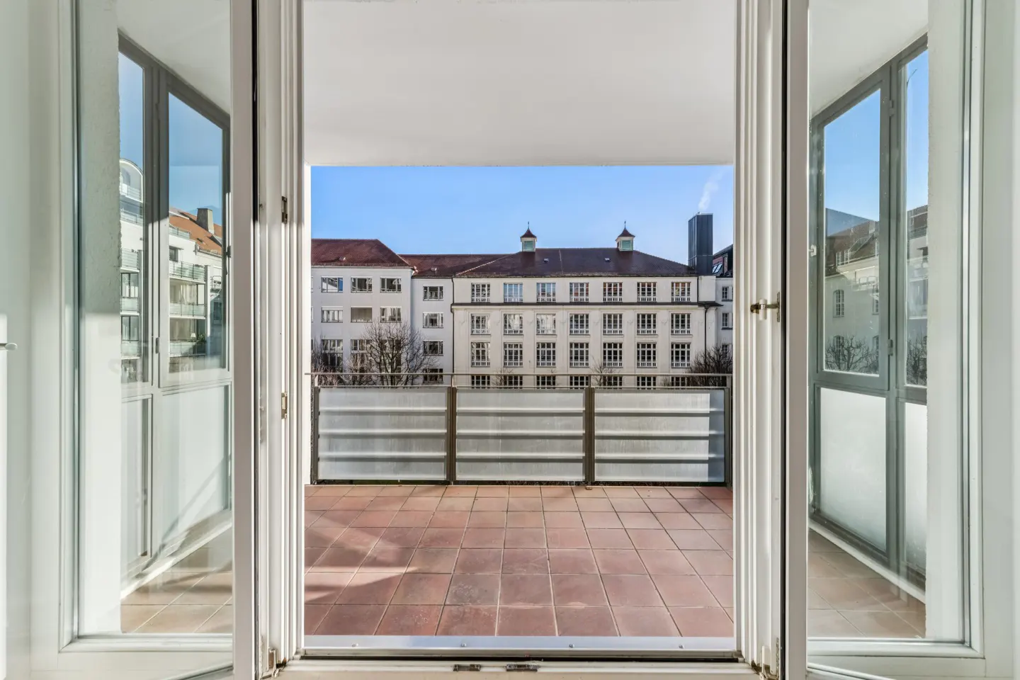 View from open white patio doors onto a terracotta tiled balcony with a metal railing and buildings in the background.
