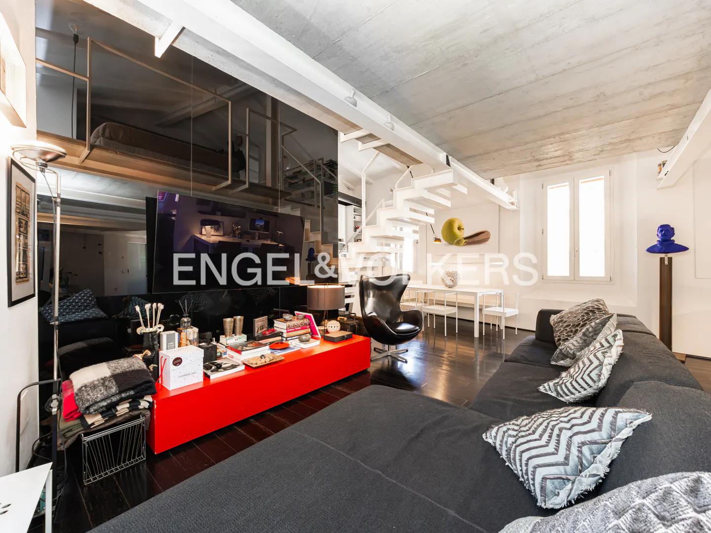 Interior view of a modern living room with a black sofa, red coffee table, and white spiral staircase. A large mirror reflects the space.