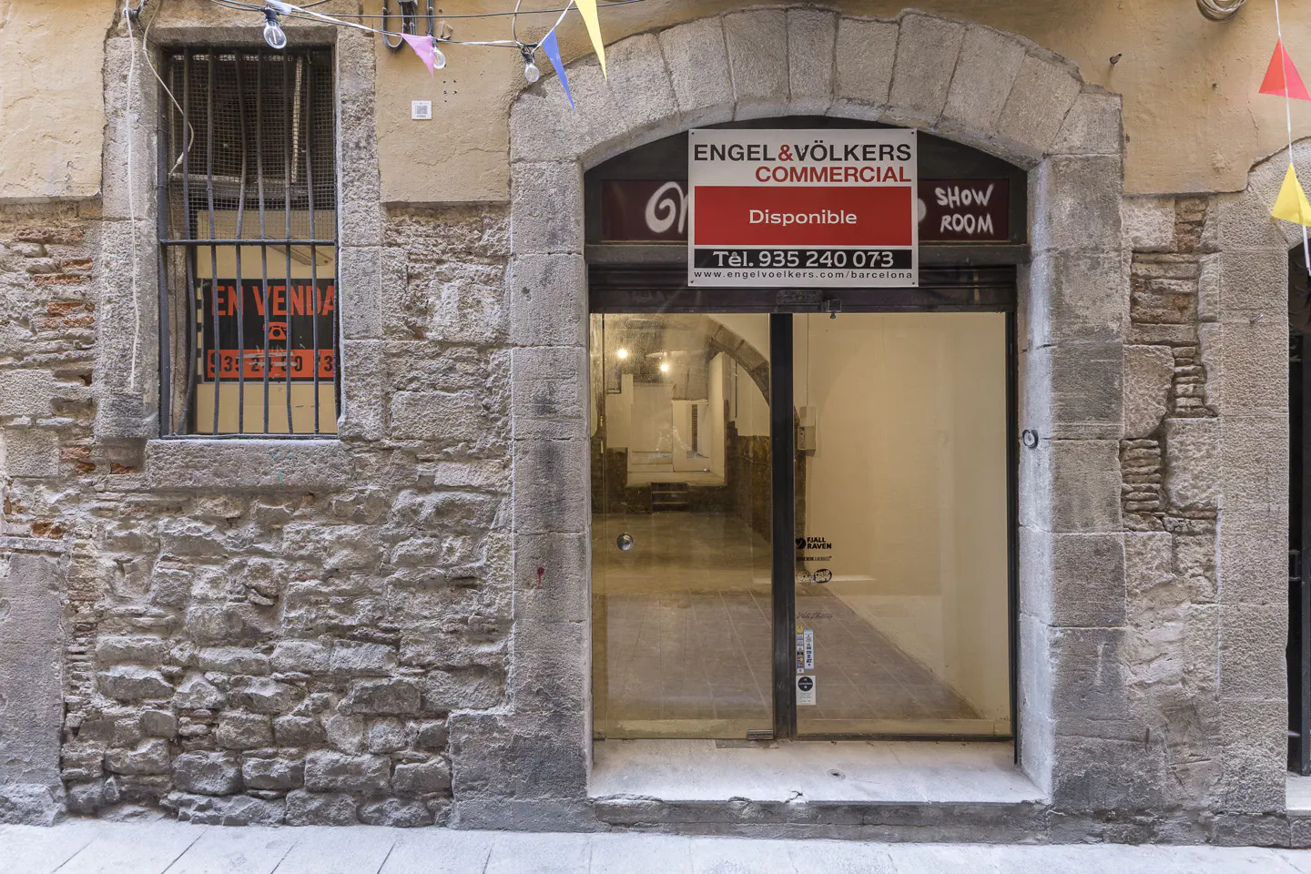 Engel & Völkers commercial space with stone facade and arched doorway. "En Venda" sign in barred window. Pennant flags hang above.