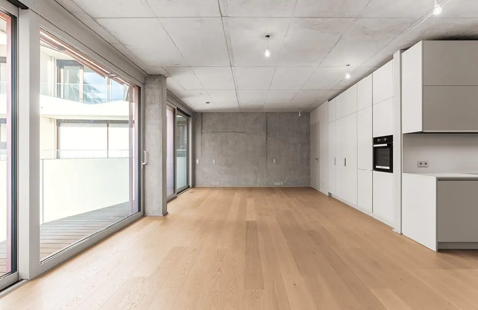 An empty, modern apartment with light wood floors, concrete walls and ceiling, and white kitchen cabinets. Sliding glass doors lead to a balcony.