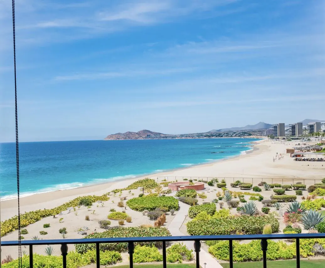 Ocean view from a balcony. Turquoise water meets a sandy beach with green vegetation. Buildings are visible in the distance under a blue sky.