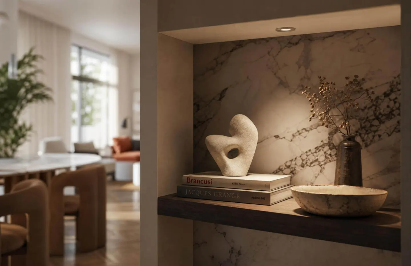 A lit marble niche displays a modern stone sculpture, art books, a bowl, and a vase, with a blurred living room beyond.