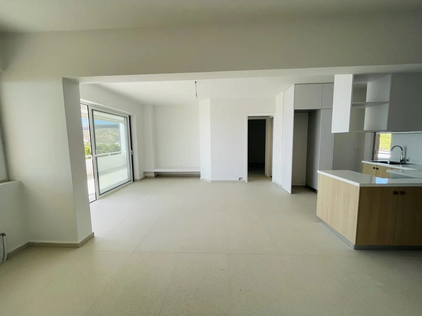 A bright, empty apartment with white walls, light tile floors, and a modern kitchen with wood cabinets. A sliding glass door leads to a balcony.