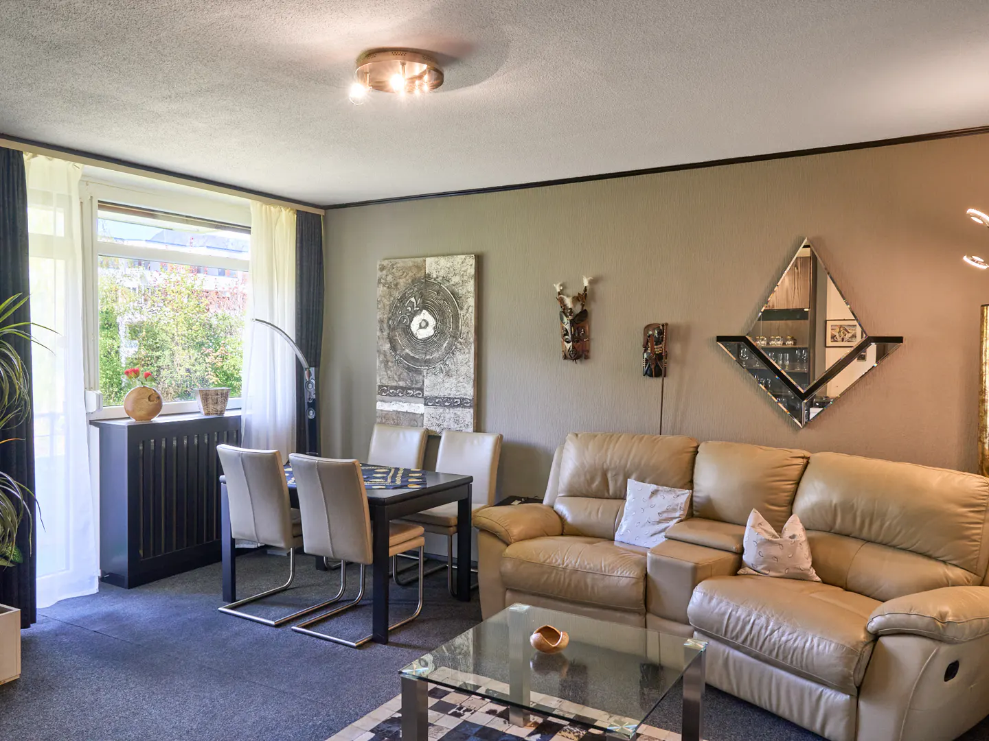 Living room with beige leather sofa, glass coffee table, and black dining table with four chairs. Artwork and a diamond-shaped mirror adorn the wall.