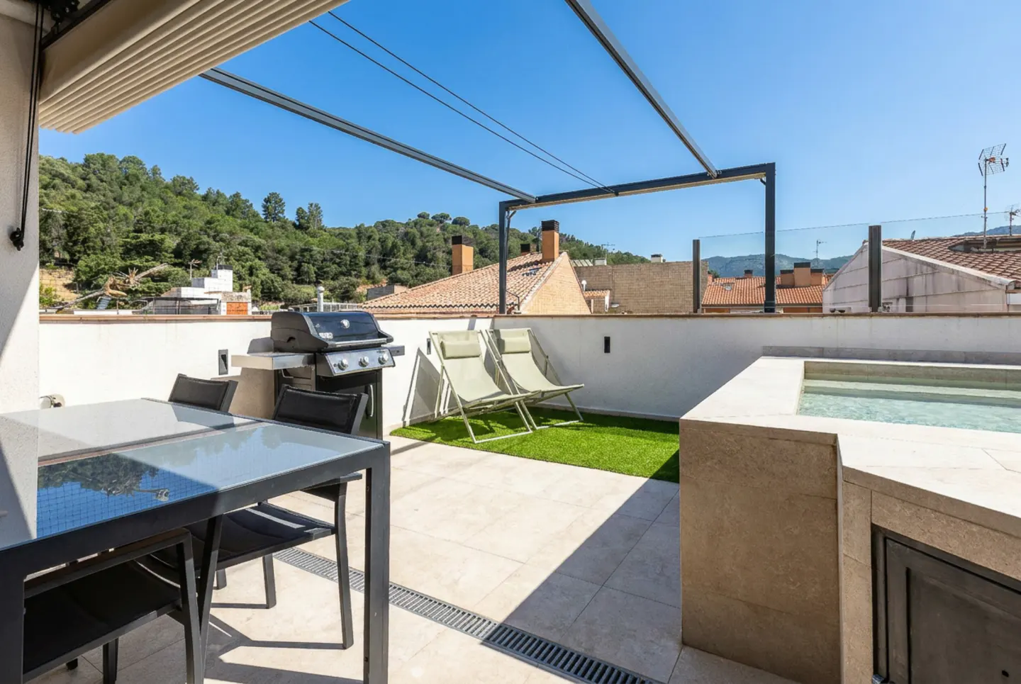 Rooftop terrace with a dining table, grill, two lounge chairs on artificial grass, and a small pool against a hill view.