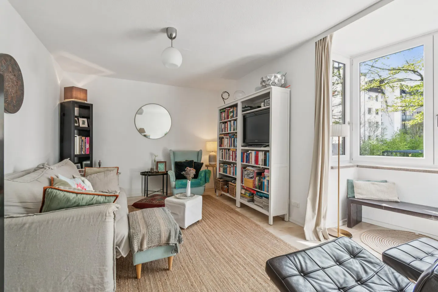 Bright living room with a beige sofa, a white bookcase filled with books, and a large window with a view of trees.