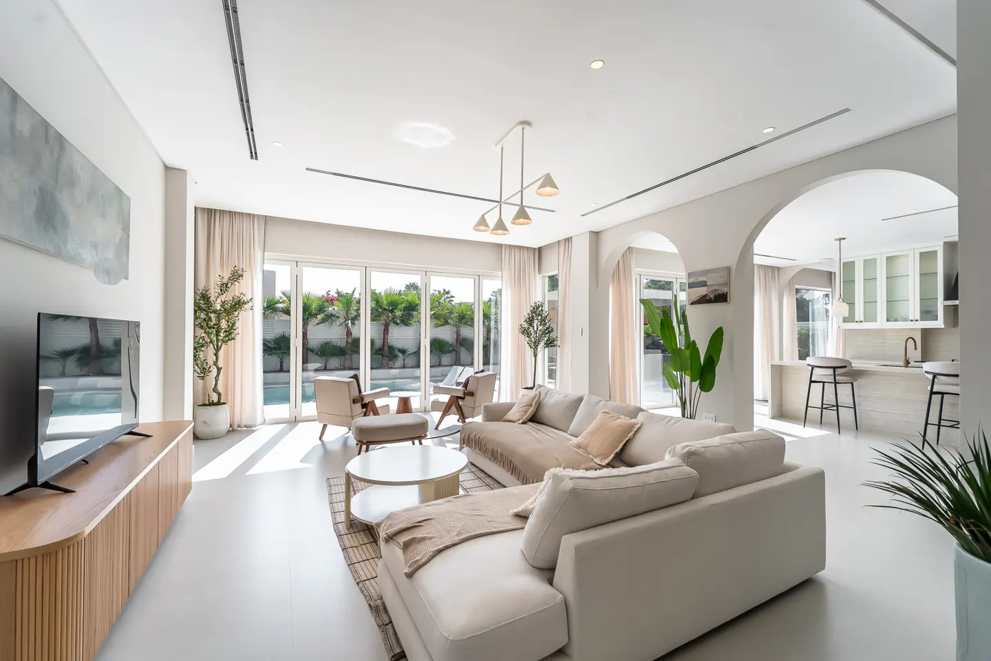 Bright, modern living room with a white sofa, chairs, and a view of the pool through sliding glass doors. A kitchen is visible through an arched doorway.