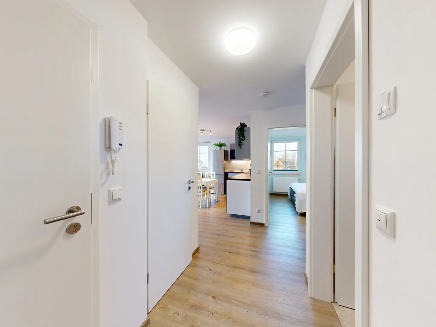 Bright hallway with light wood floors, white walls, and doors. A kitchen and bedroom are visible in the background.