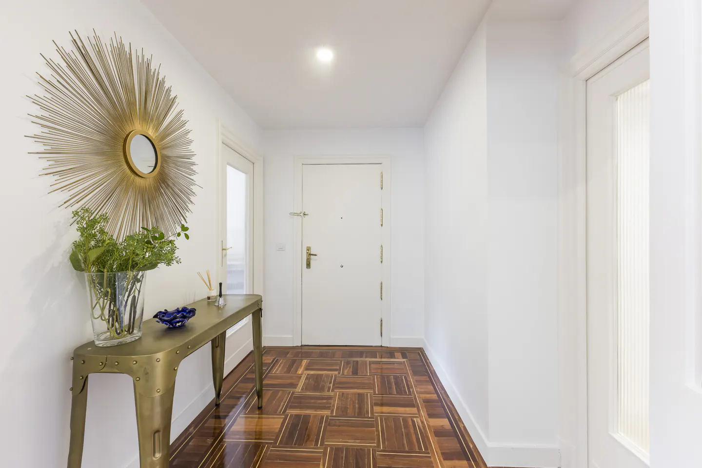 Hallway with parquet floor, white walls, and a gold console table with a sunburst mirror above it.