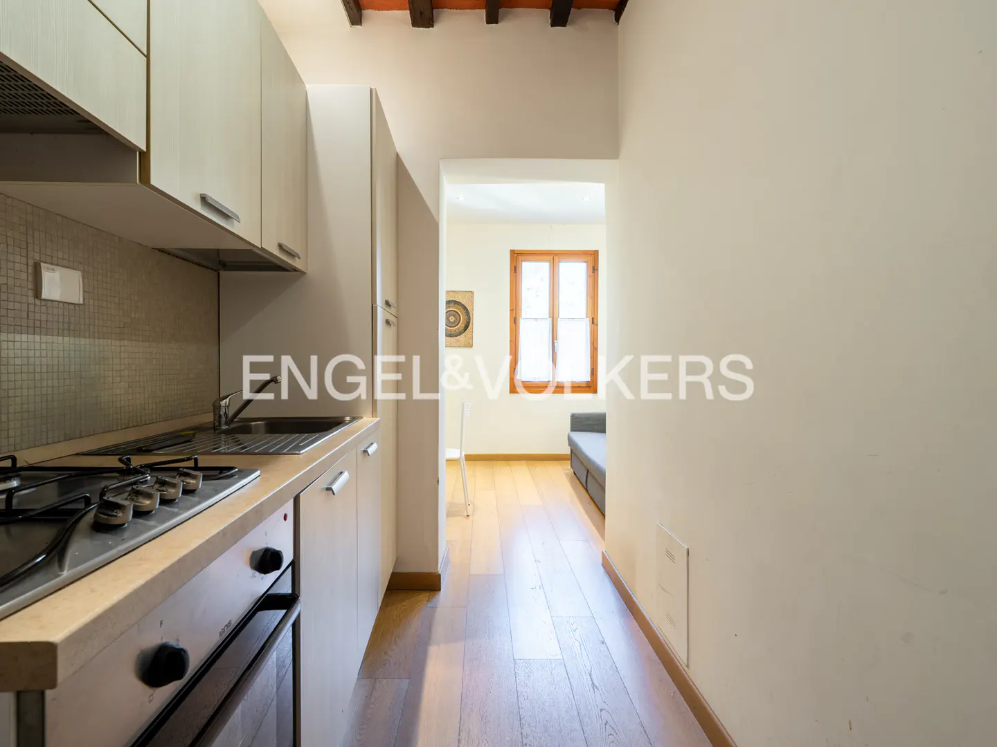 Modern kitchen with light wood cabinets, stainless steel stove, and hardwood floors leading to a bright living area.