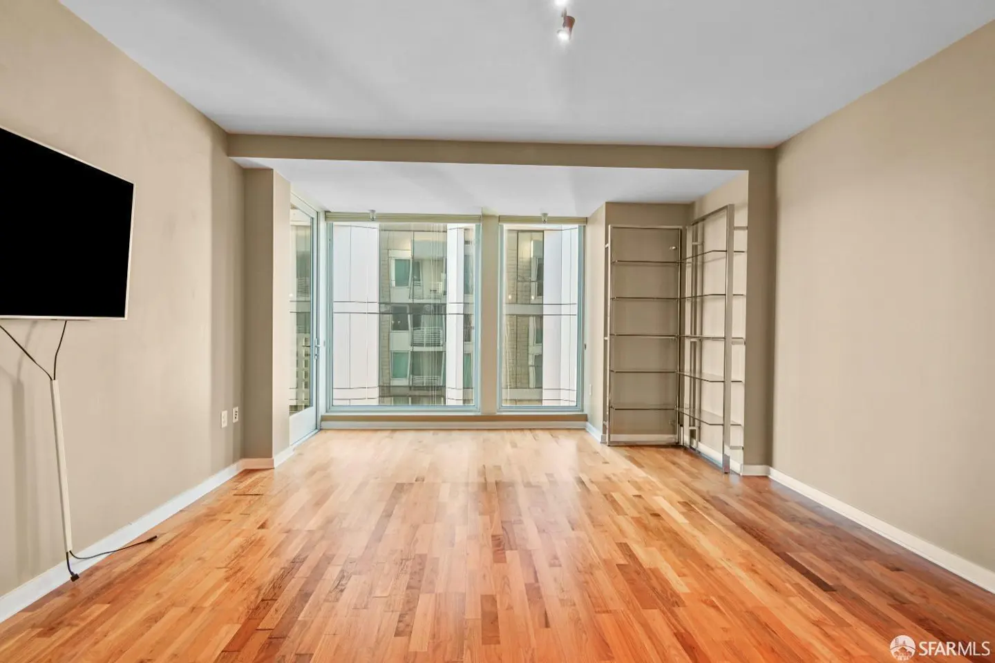 A bright, empty room with hardwood floors, beige walls, and large windows showing a city view. A TV is mounted on the left wall.