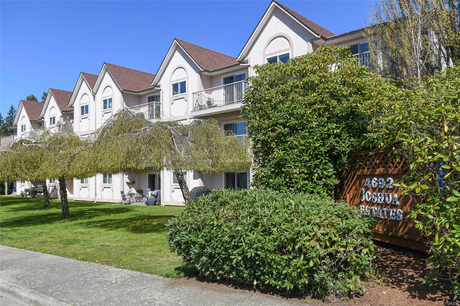 Exterior view of Joshua Estates, a multi-story apartment building with brown roofs and white walls, surrounded by green trees and bushes.