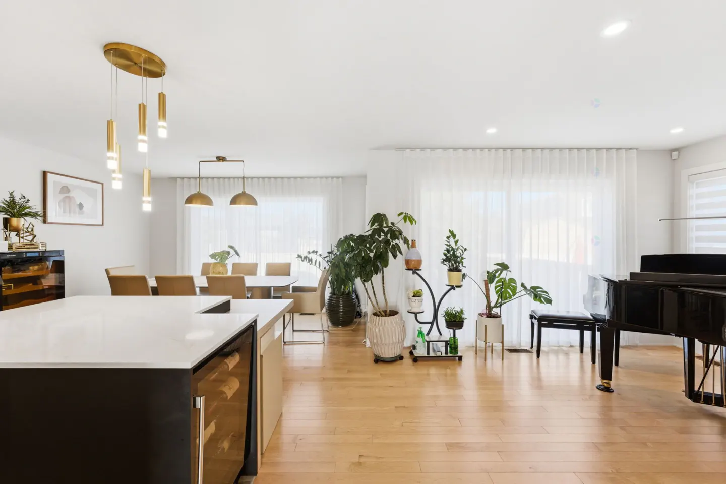 Bright, open-concept living space with a black island, dining table, plants, and a black grand piano on a light wood floor.