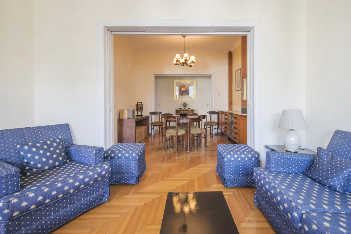 Living room with blue patterned sofas, ottomans, and a black coffee table. A dining room with a table and chairs is visible through a doorway.