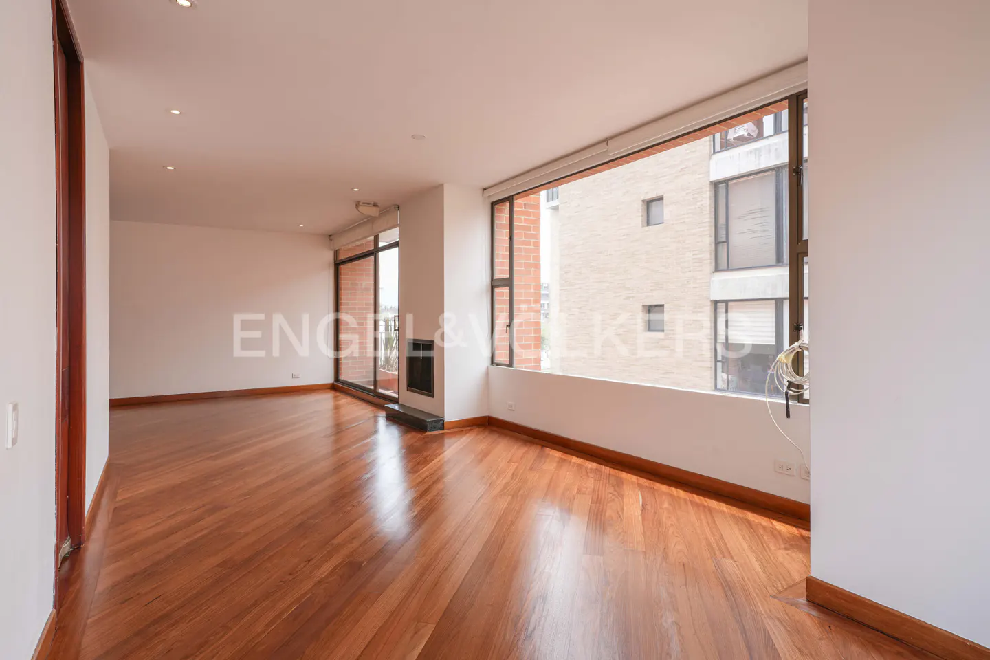 Empty living room with polished hardwood floors, white walls, a fireplace, and large windows overlooking city buildings.