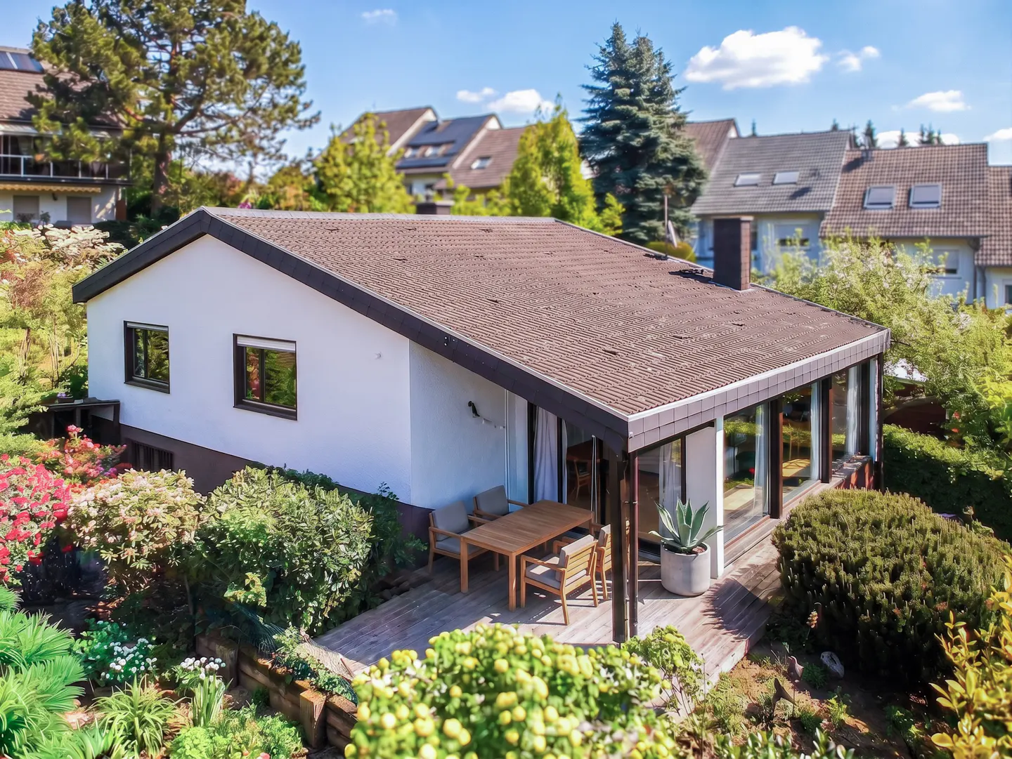 Exterior view of a white house with a brown roof, surrounded by lush greenery and a wooden deck with outdoor furniture.