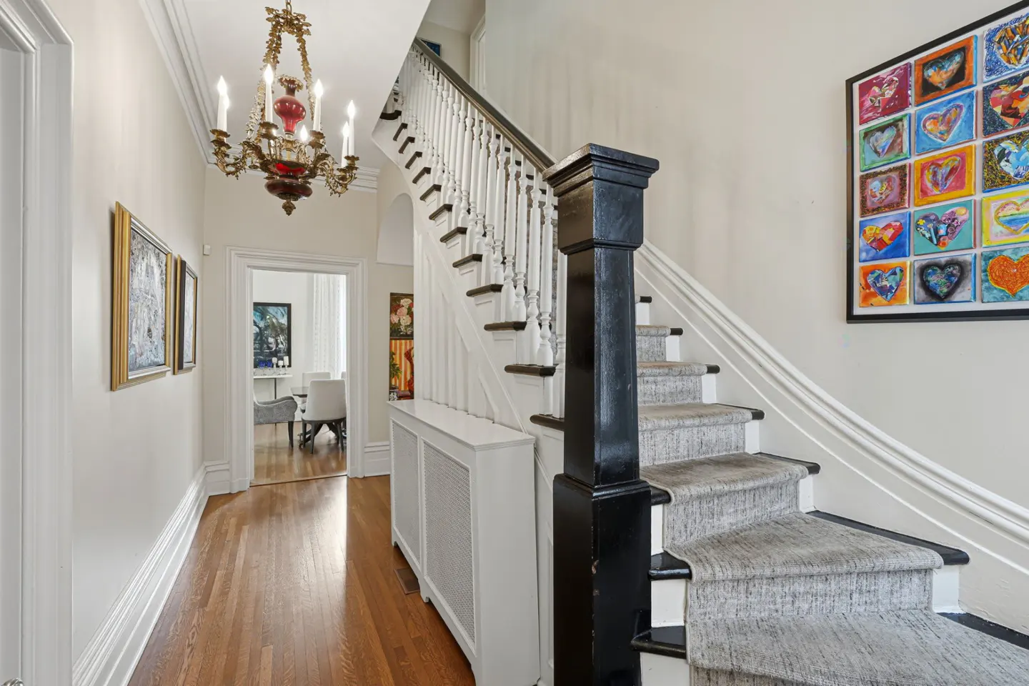 Foyer with hardwood floors, white walls, and a staircase with black banister and gray carpet. A colorful heart art piece hangs on the wall.