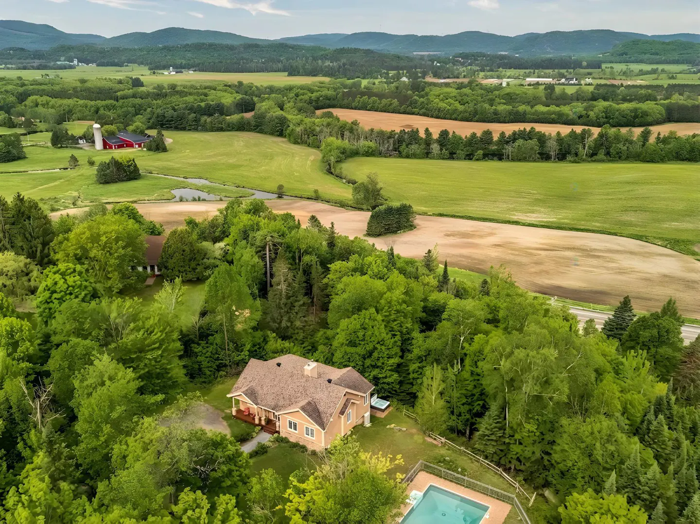 Aerial view of a tan house with a pool, nestled in lush trees overlooking vast green fields and distant mountains.