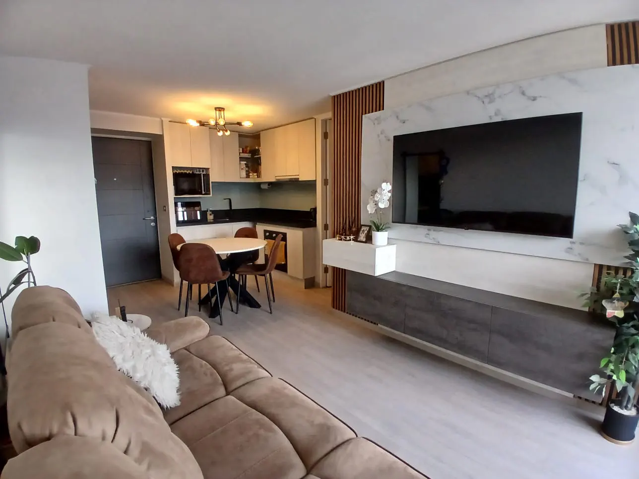 A modern apartment interior with a brown sofa, round dining table, and a large TV mounted on a marble wall.