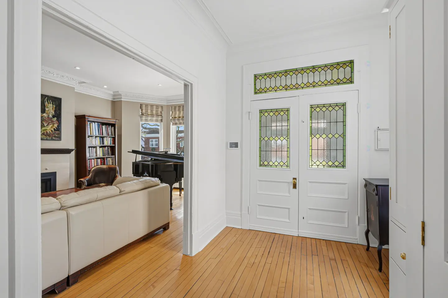 Bright foyer with hardwood floors, white walls, and double doors with stained glass. Living room visible with piano, sofa, and bookcase.