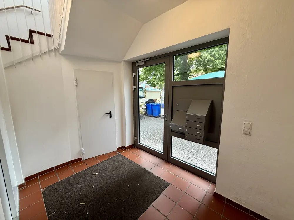 Bright apartment building entrance with tiled floor, a dark doormat, a white door, and a glass door with mailboxes.