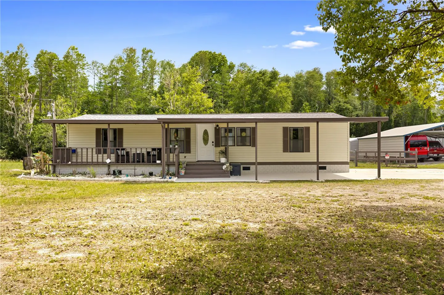 Tan single-story home with a covered porch and brown trim, set on a grassy lawn with trees in the background.
