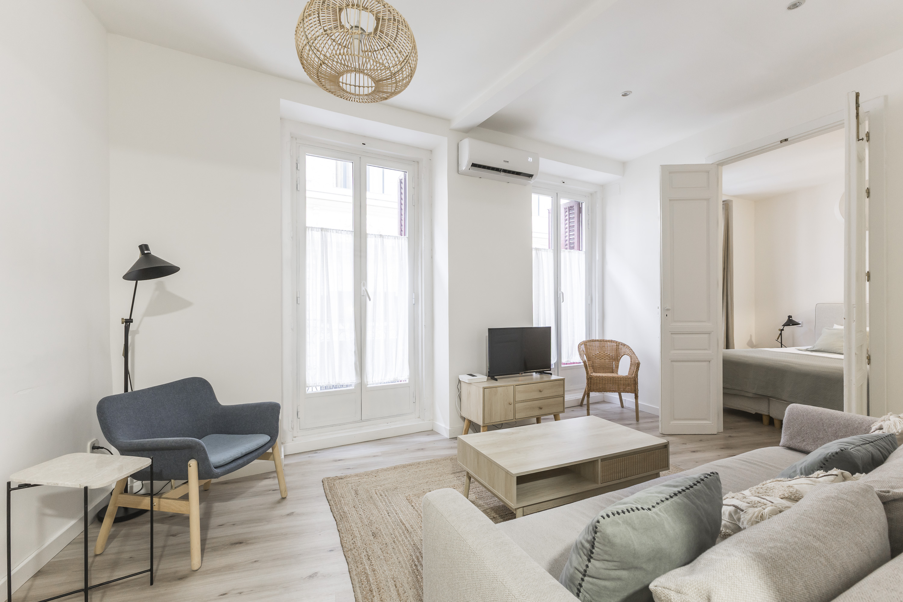 Bright living room with a grey sofa, blue armchair, wooden coffee table, and a view into a bedroom through open doors.