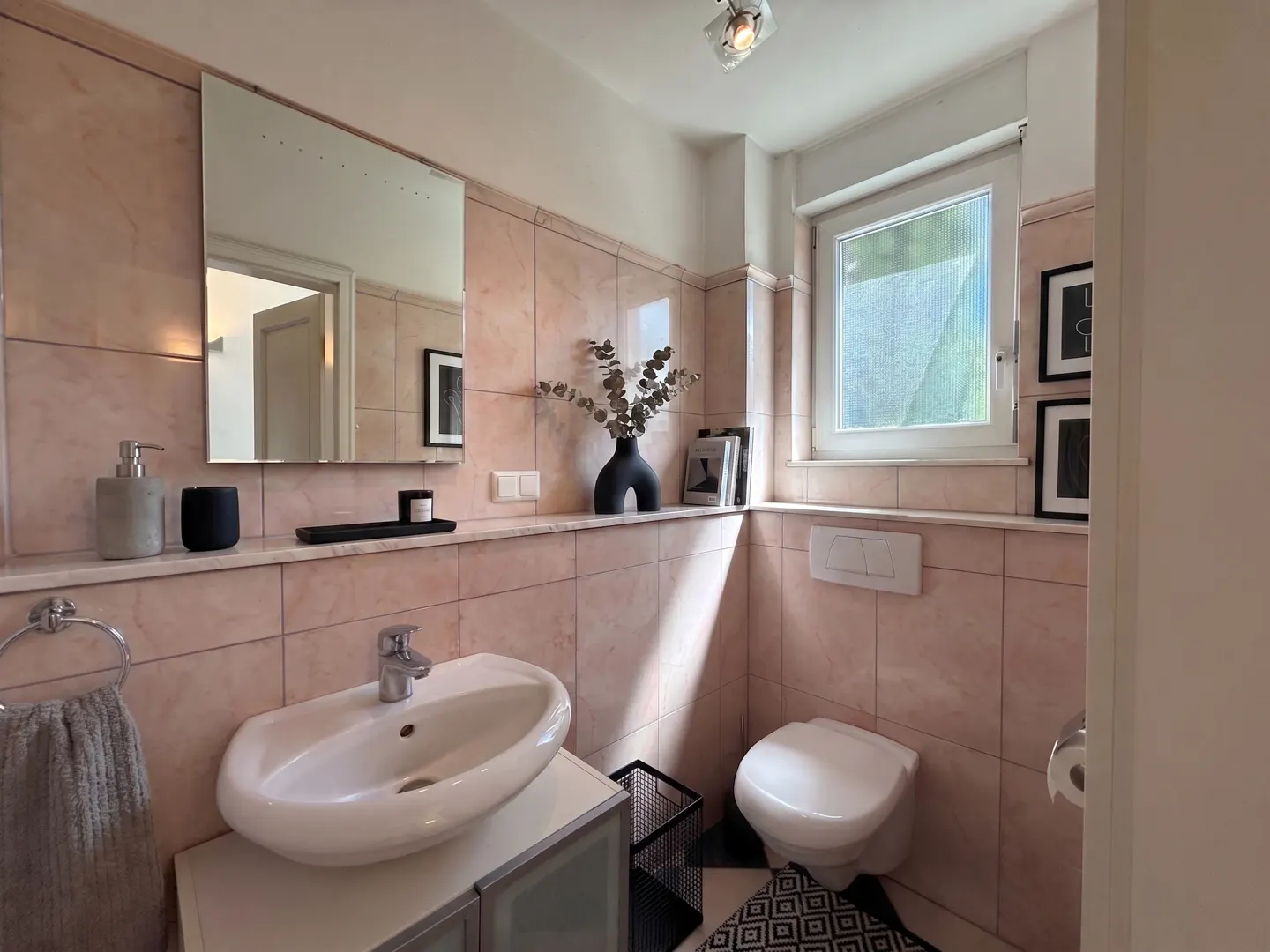 Bathroom with pink tile walls, white sink and toilet, black and white patterned floor, and a window.