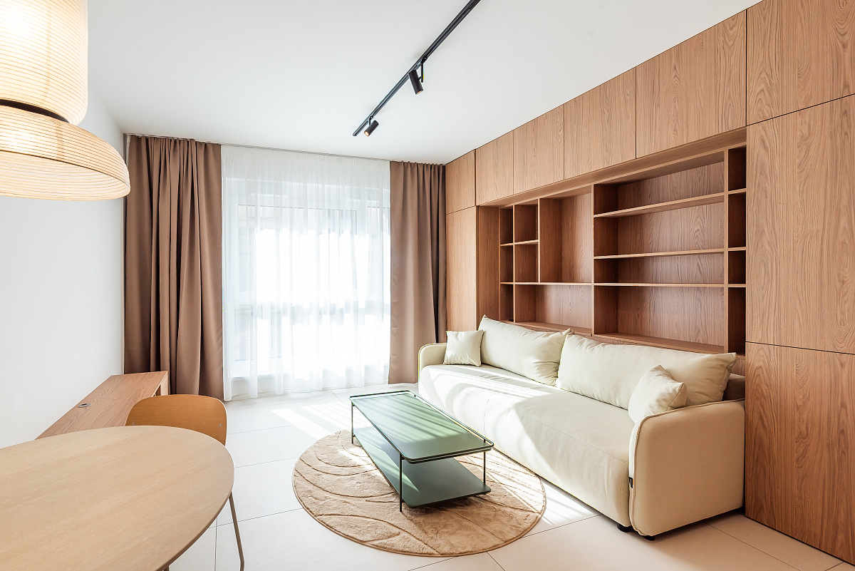 Modern living room with a cream sofa, green coffee table on a round rug, and a large wooden wall unit with built-in shelves.