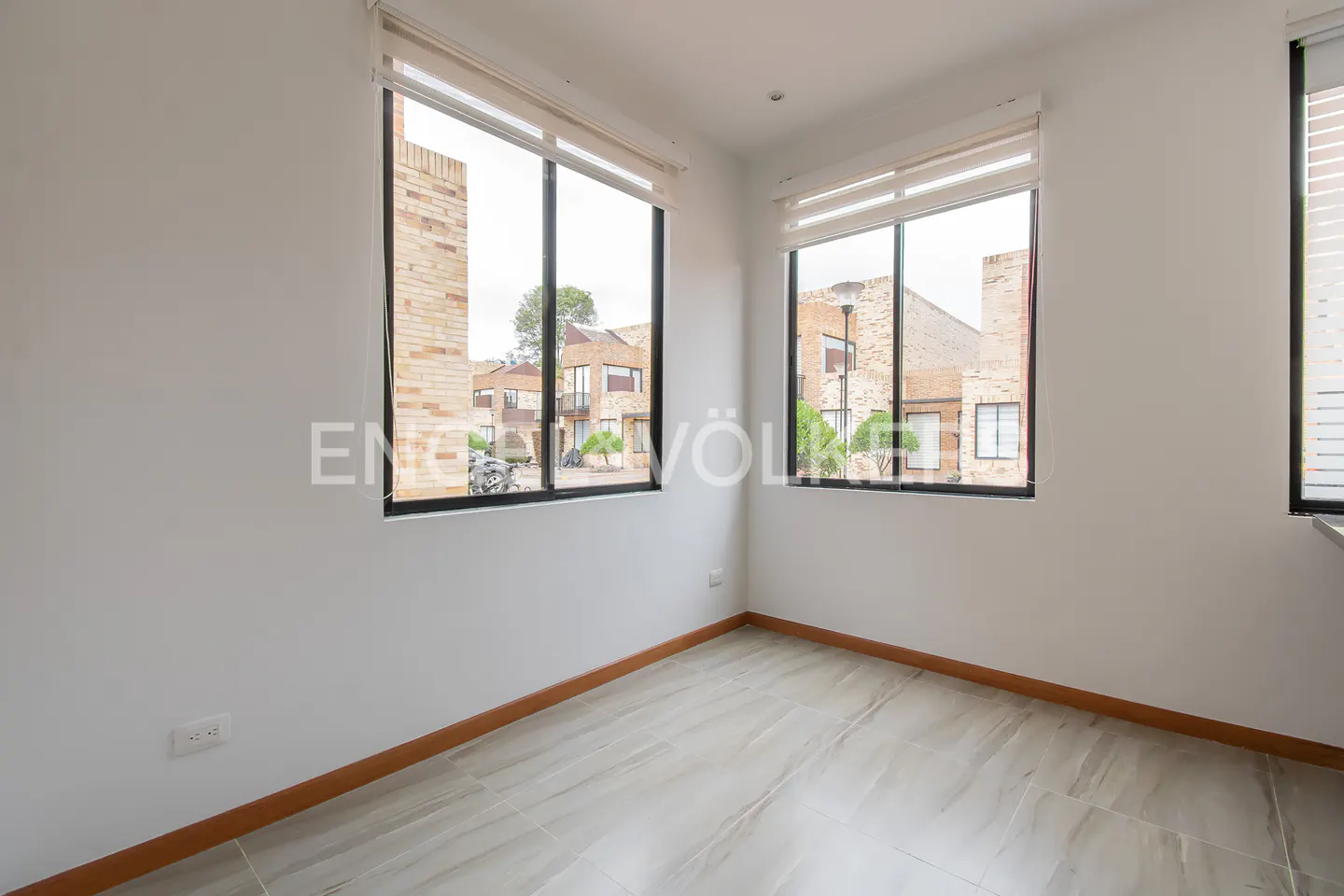 Empty room with white walls, gray tile floor, and two large windows with black frames and white blinds. Outside are brick buildings.
