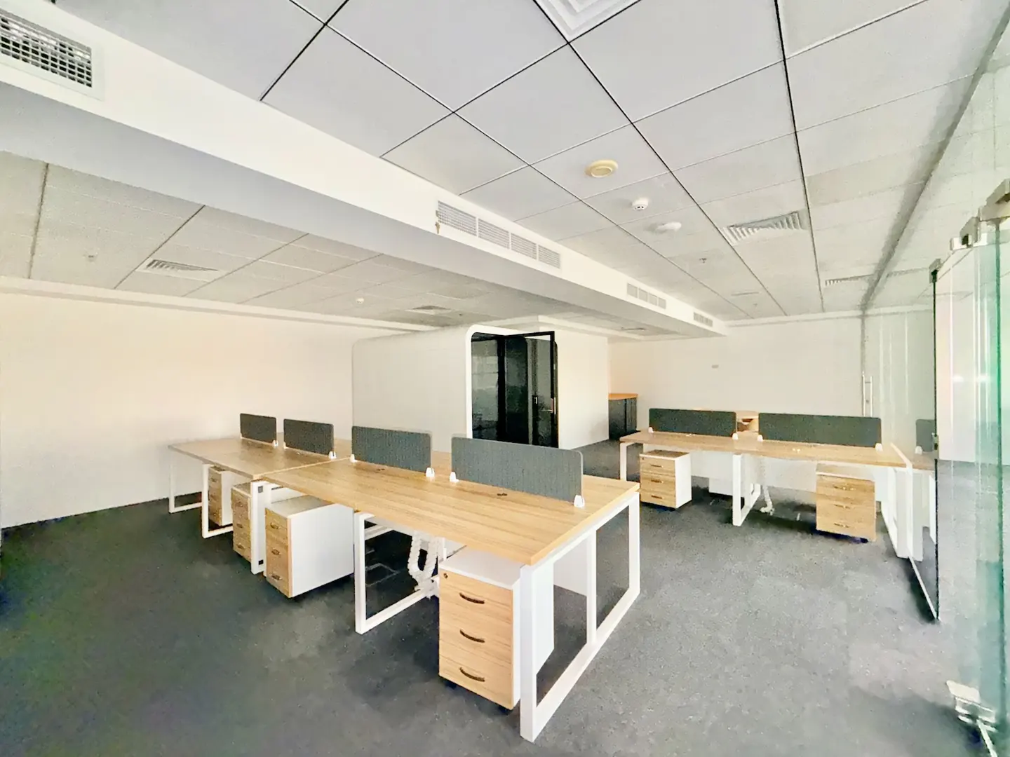 Bright office space with rows of desks, light wood tops, white frames, and gray dividers. White walls and ceiling with dark gray flooring.