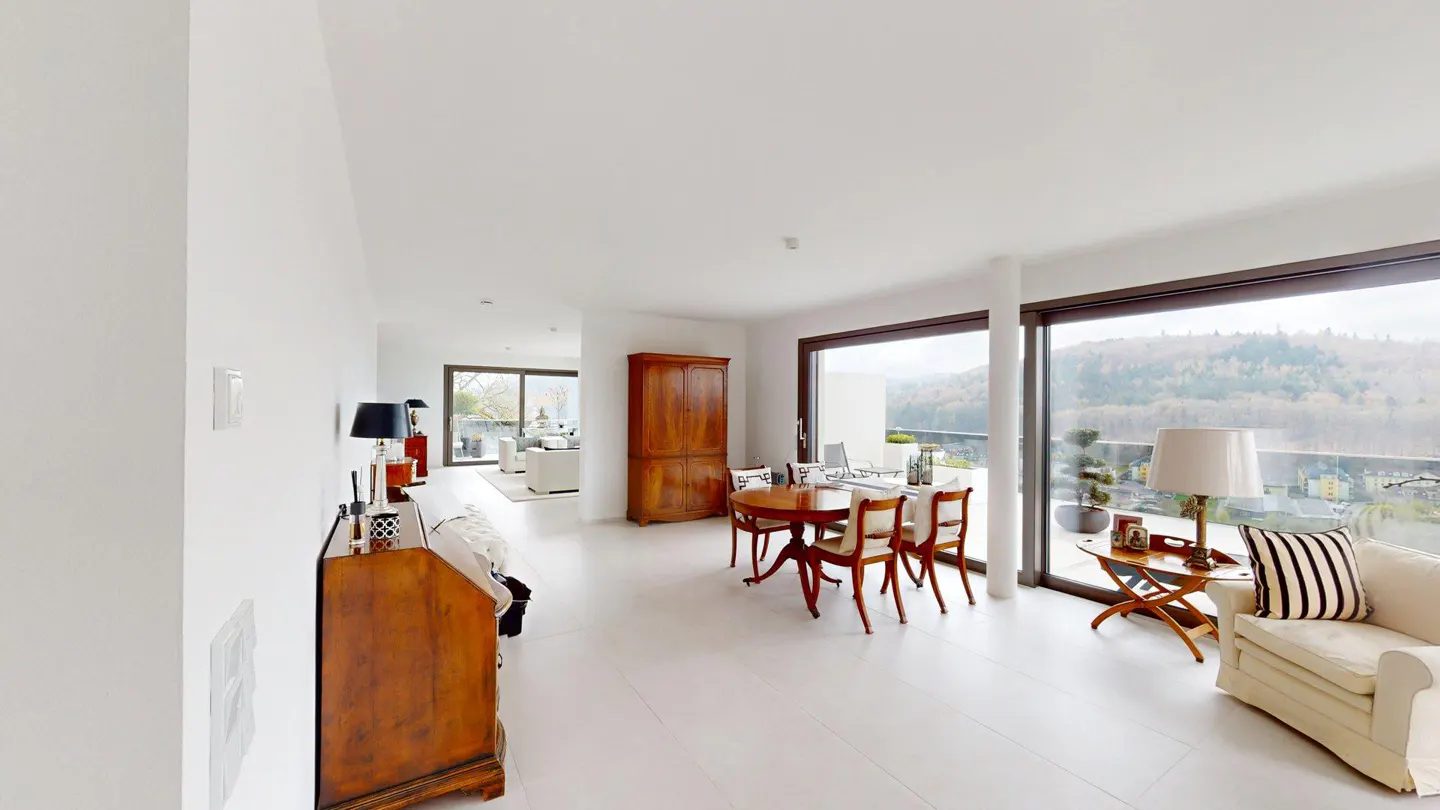 Bright, modern living room with white walls and floors. Large windows offer a view of trees. A wooden table and chairs sit near a wooden cabinet.
