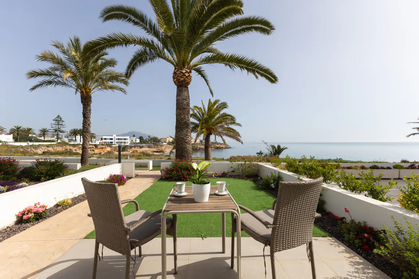 Outdoor patio with table, chairs, and ocean view. Palm trees and flowers surround the space. Two cups sit on the table.