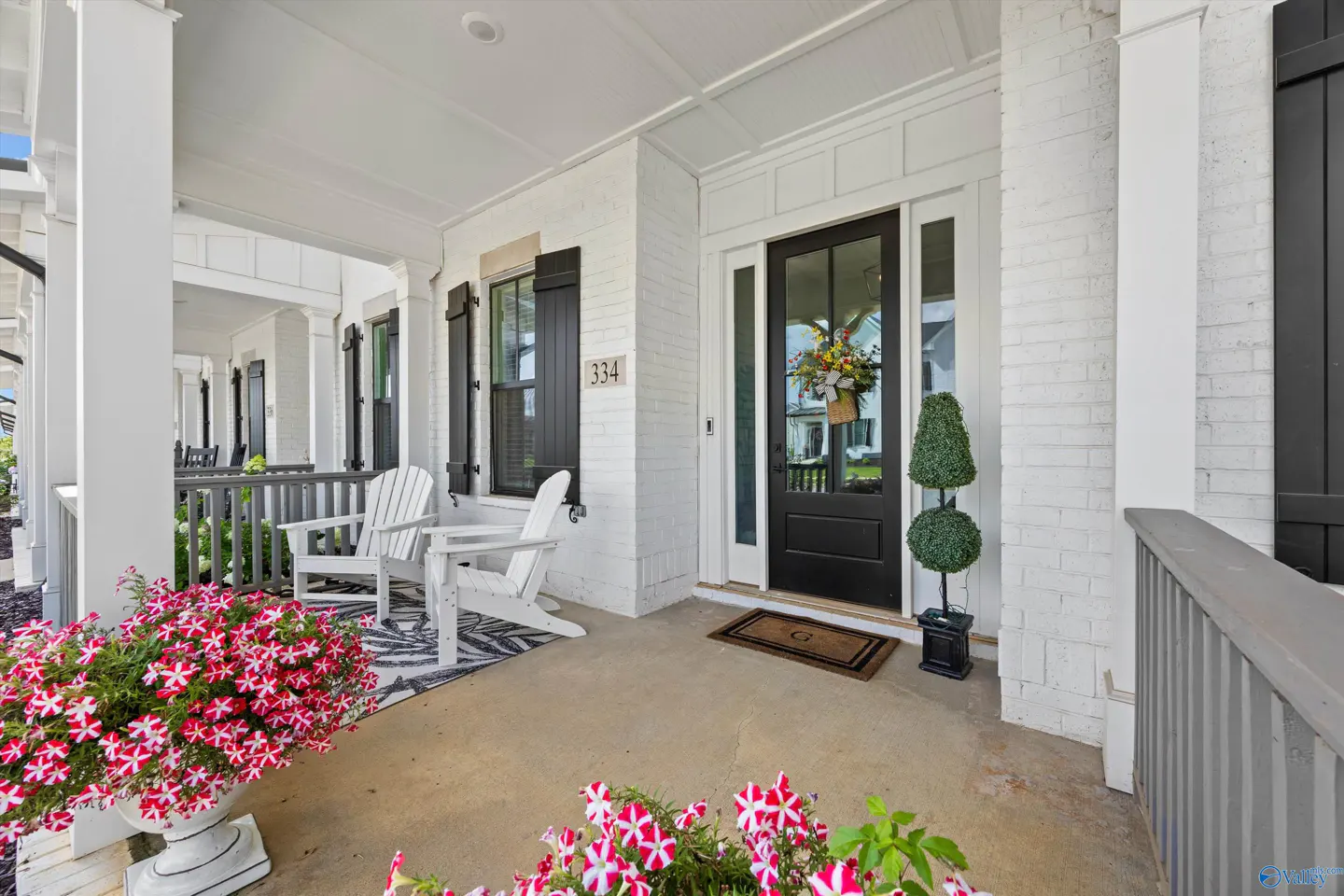 Front porch of a white brick house with black shutters, a black front door with a wreath, and two white Adirondack chairs.