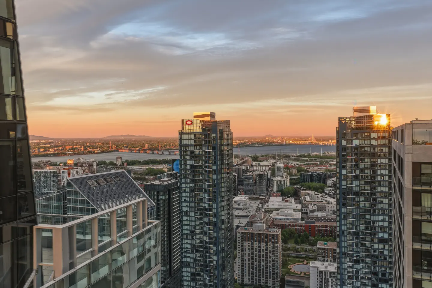 Cityscape view at sunset with tall buildings, a river, and a bridge in the background. The sky is a mix of orange, pink, and gray.