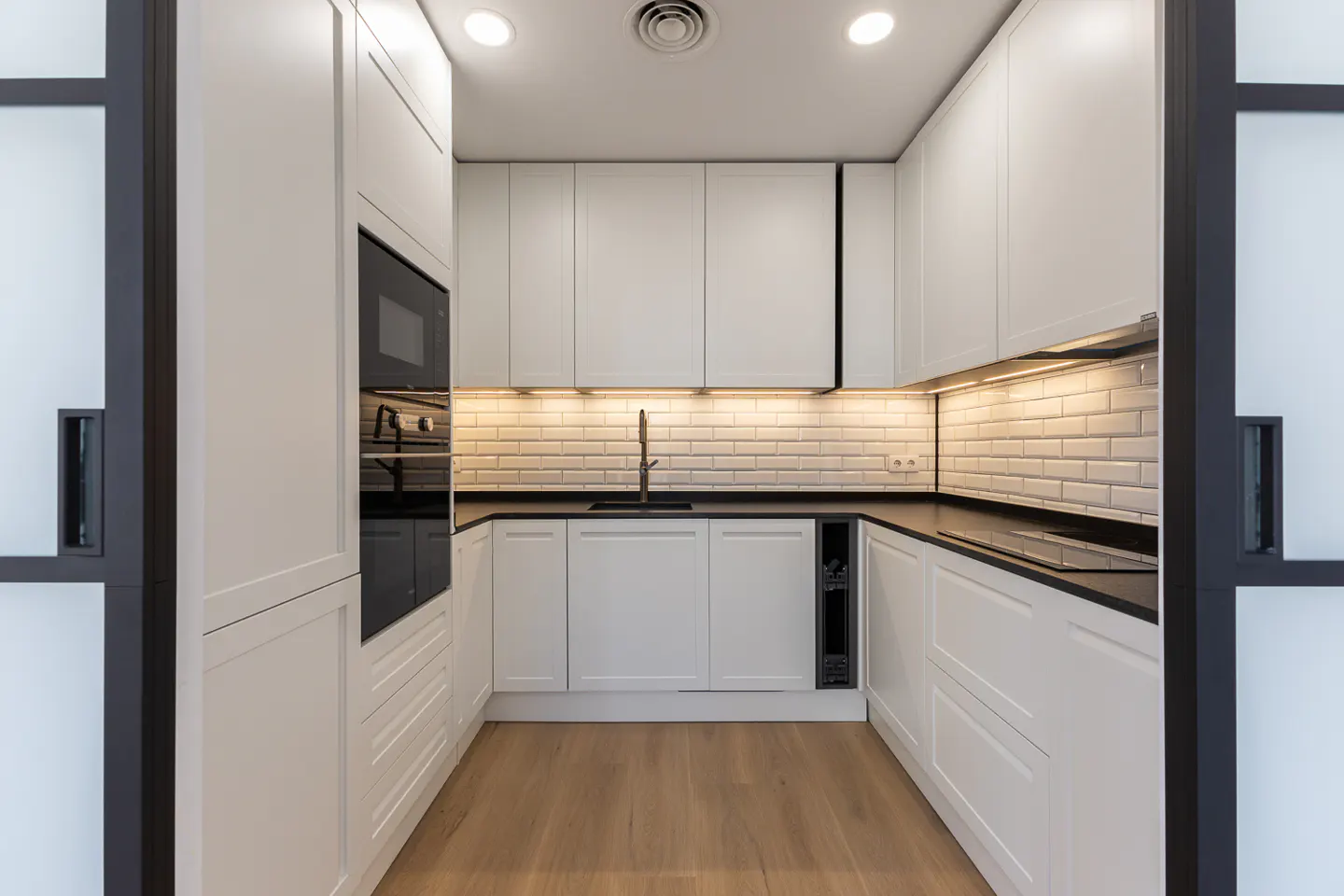 A modern kitchen with white cabinets, black countertops, and a light wood floor. Subway tile backsplash and black appliances.