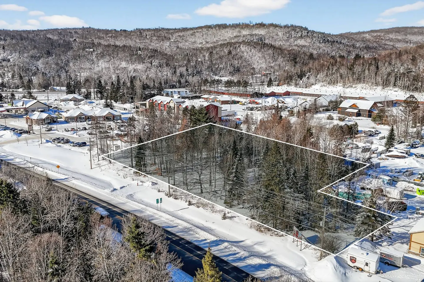 Aerial view of a snow-covered lot outlined in white, with trees, a road, and buildings in the background.