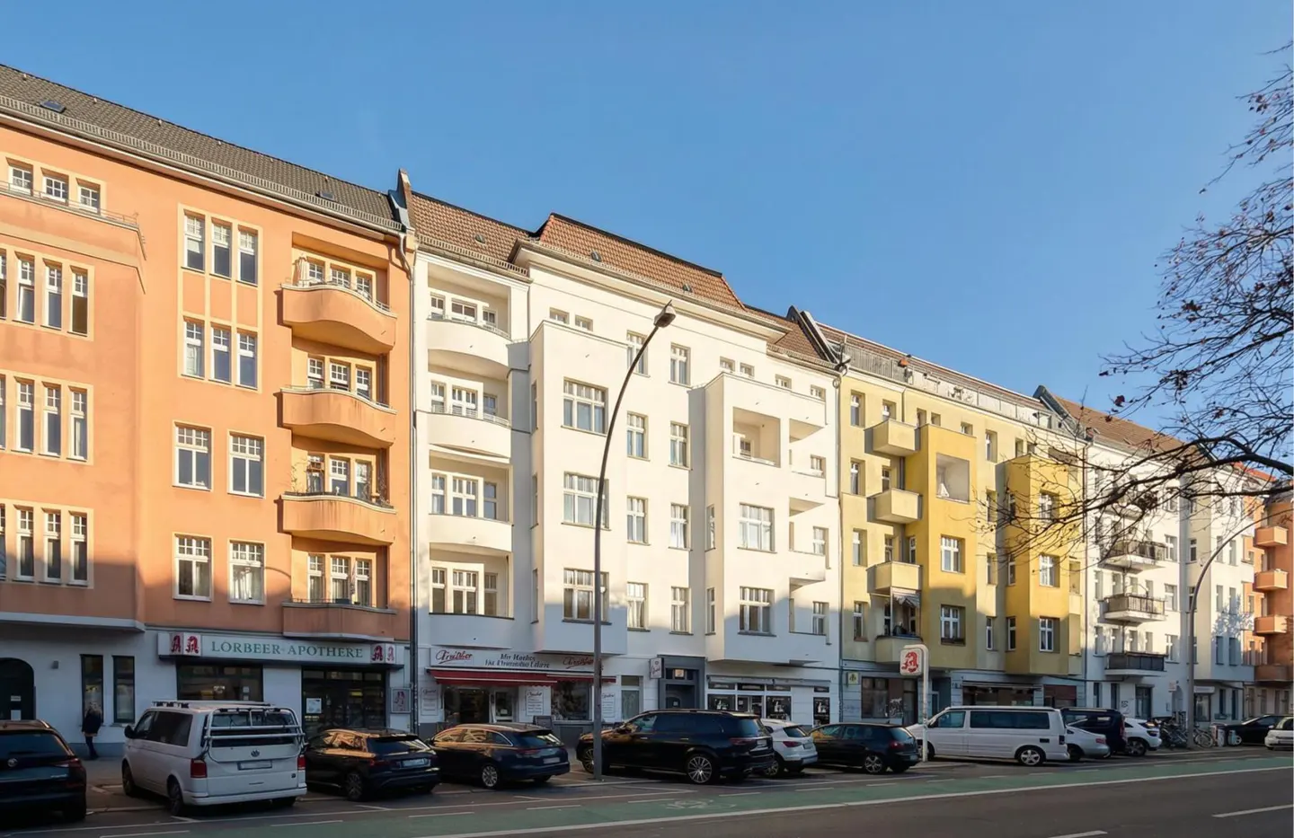 Street view of colorful apartment buildings with parked cars and shops on the ground floor under a clear blue sky.