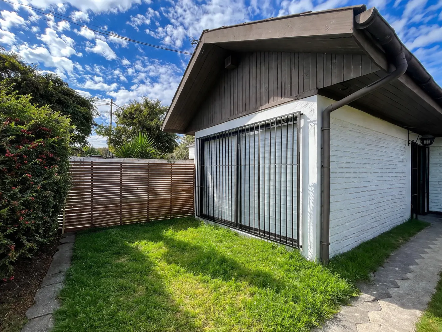 Exterior view of a white brick house with a brown roof, a barred window, and a wooden fence, under a blue sky.