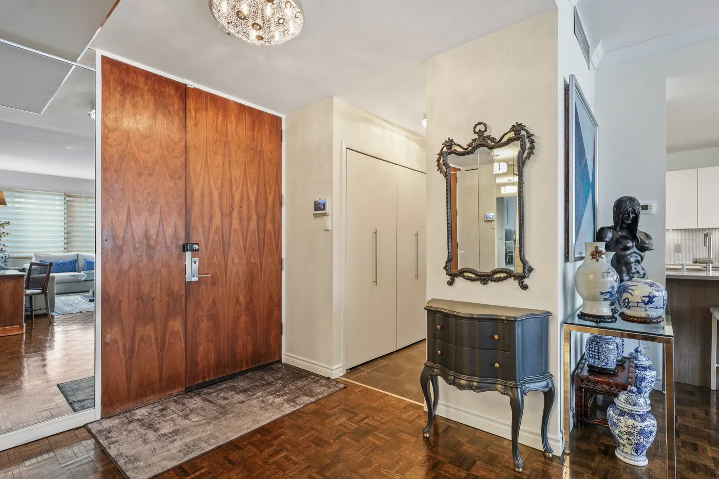 Condo foyer with wood parquet floor, large wood door, and mirrored wall. A black chest with mirror and glass table with vases decorate the space.