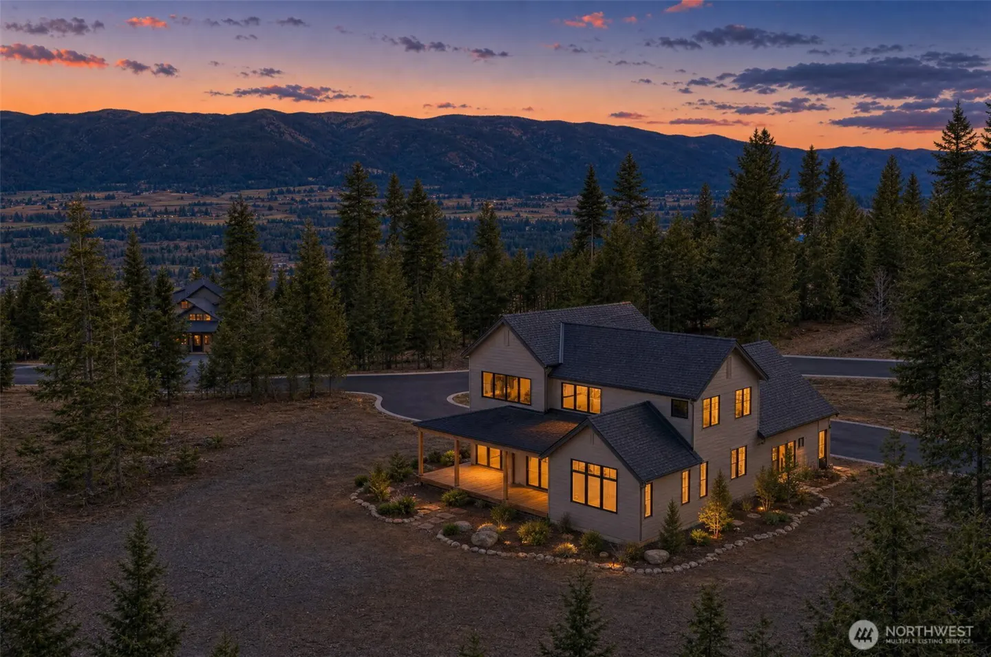 A two-story house with a dark roof sits among trees at dusk, lights glowing from the windows. Mountains are visible in the background.