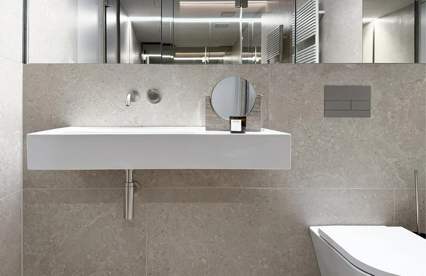 Modern bathroom featuring a white, rectangular sink, round mirror, and gray marble walls. A toilet is partially visible on the right.