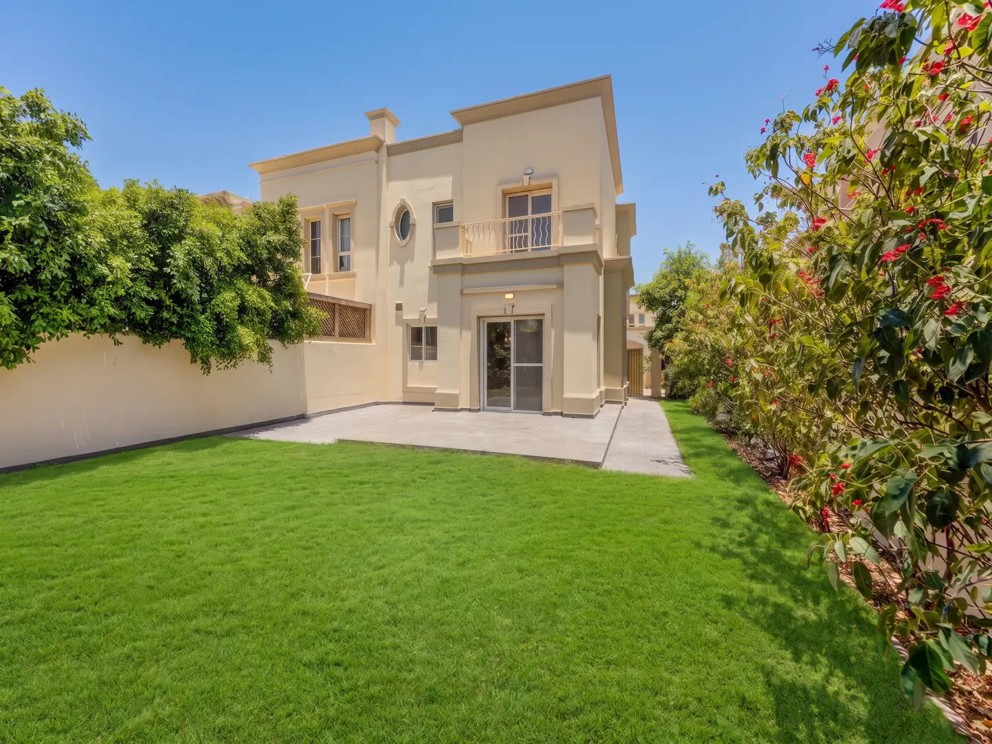 A two-story beige house with a balcony and sliding glass door, surrounded by green grass and trees under a blue sky.