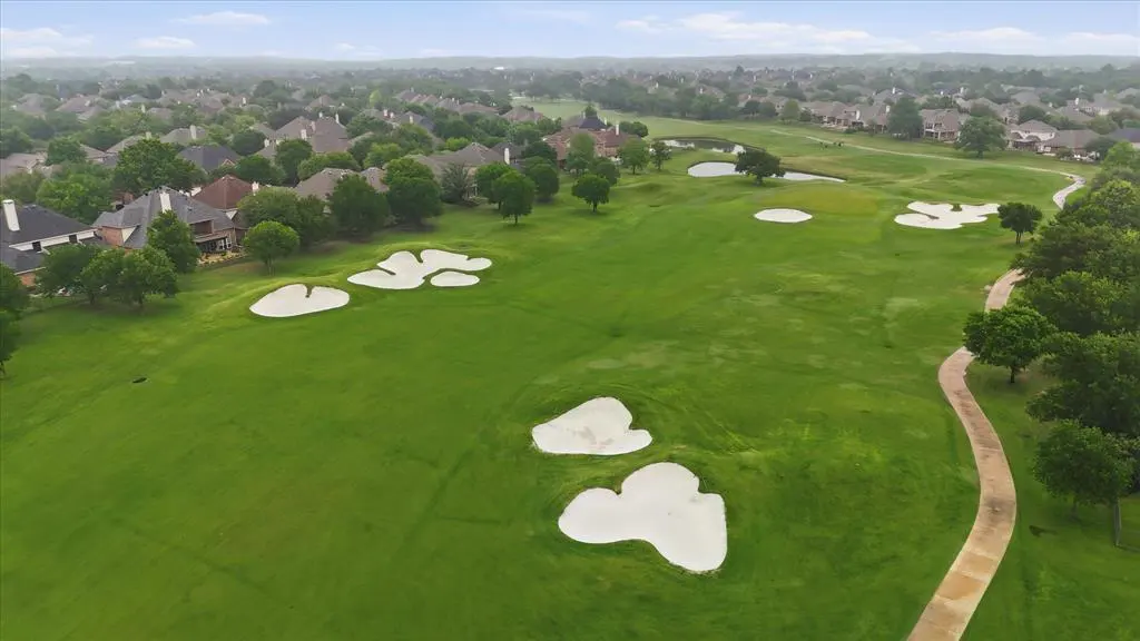 Aerial view of a lush green golf course with white sand traps, a winding path, and houses in the background.