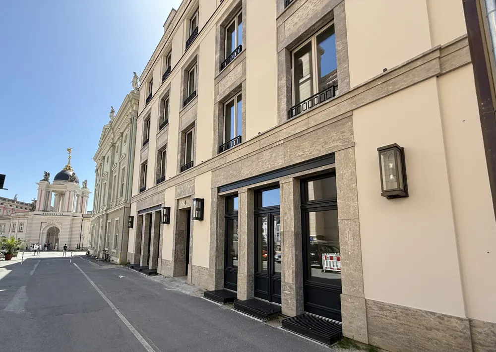 Modern beige building with stone trim and black-framed windows on a street near a historic domed building.