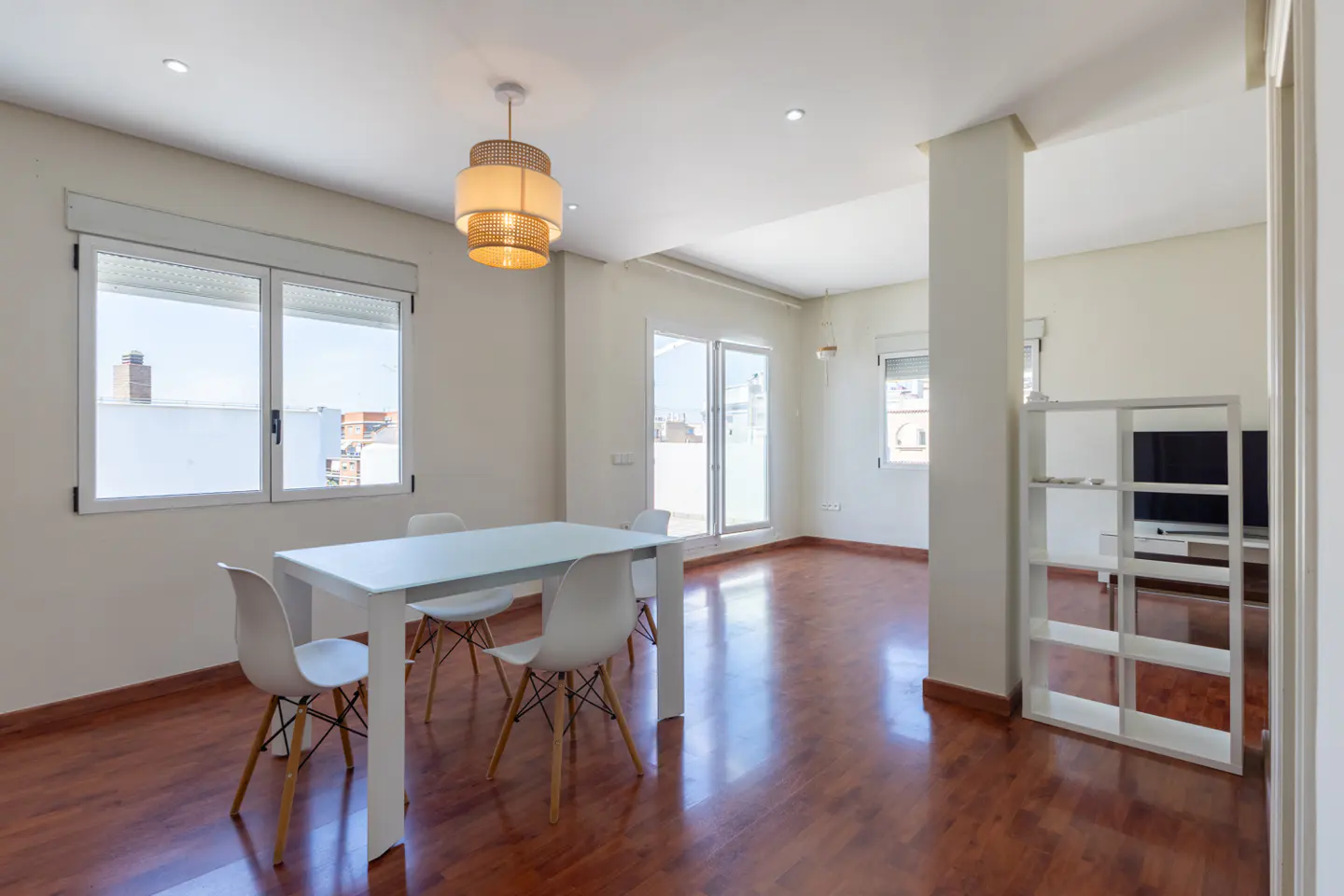 Bright, open-plan apartment with a white table and chairs on a wood floor, lit by a woven pendant light. Windows overlook a cityscape.