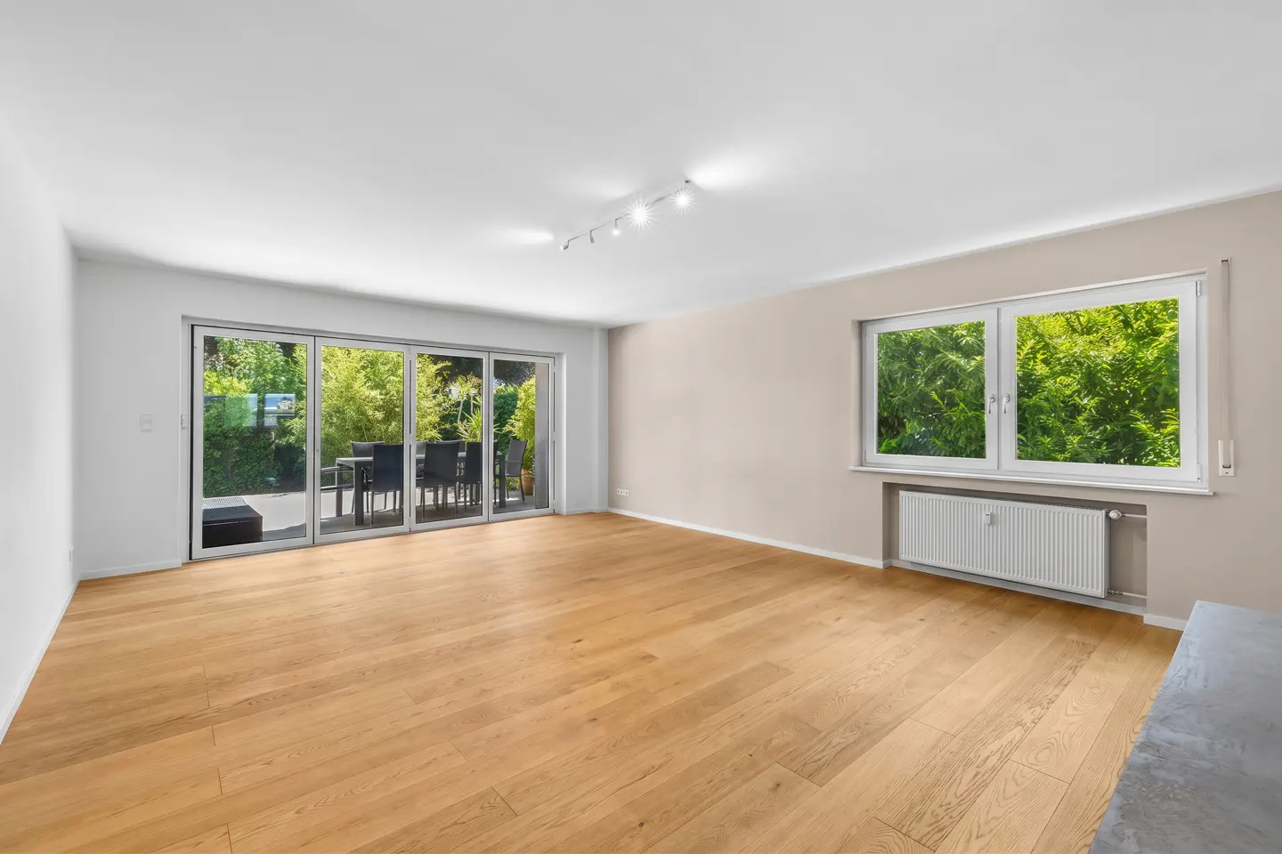 Spacious, empty living room with light wood floors, beige walls, large glass patio doors, and a window with garden views.