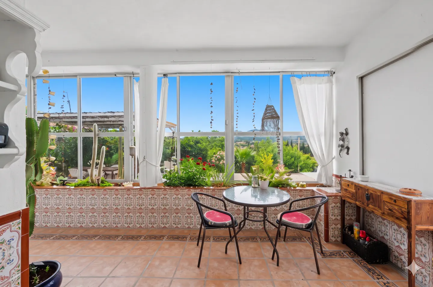 Bright sunroom with tiled floor, large windows, and garden view. A small table and two chairs sit in the center.