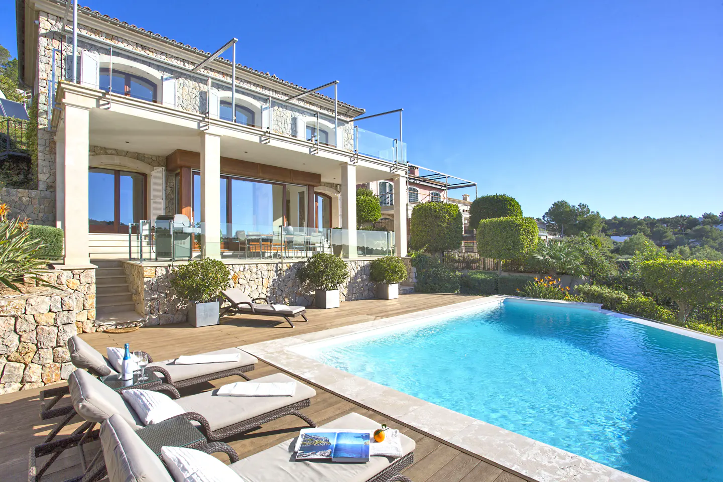 Luxury home exterior with a pool, lounge chairs, and a stone facade under a clear blue sky.